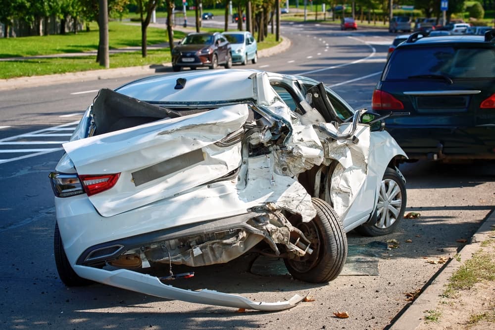 A heavily damaged white car on the side of the road after a collision, showing severe rear and side impact damage, representing property damage from a negligent driver.