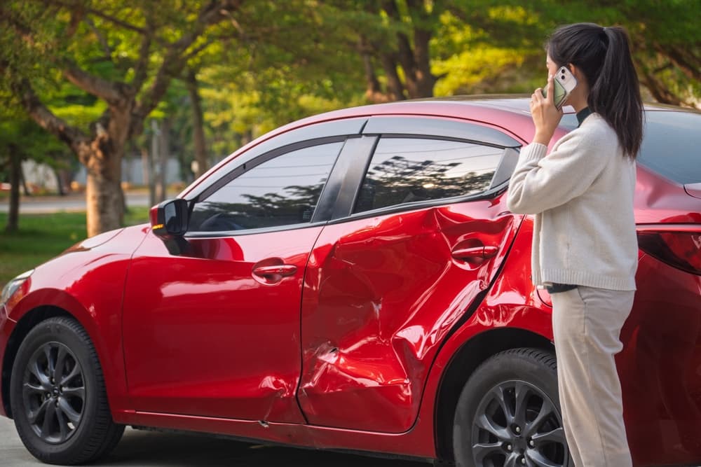 Woman on phone standing beside a red car with severe side damage after a collision in a park area