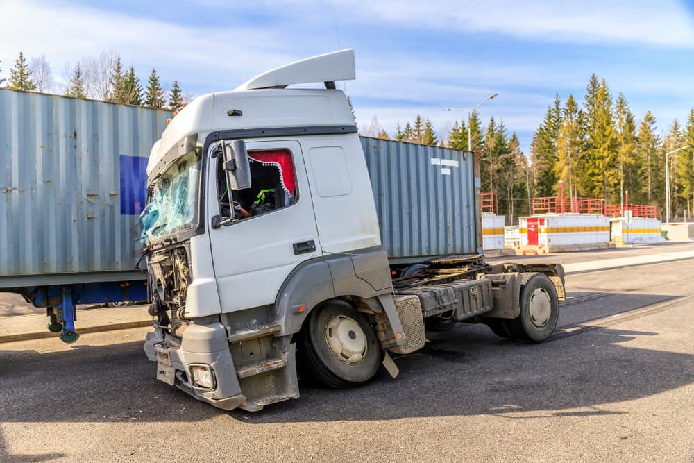 Damaged semi-truck cab jackknifed against its trailer after an accident on a road, with a shattered windshield and bent front bumper.
