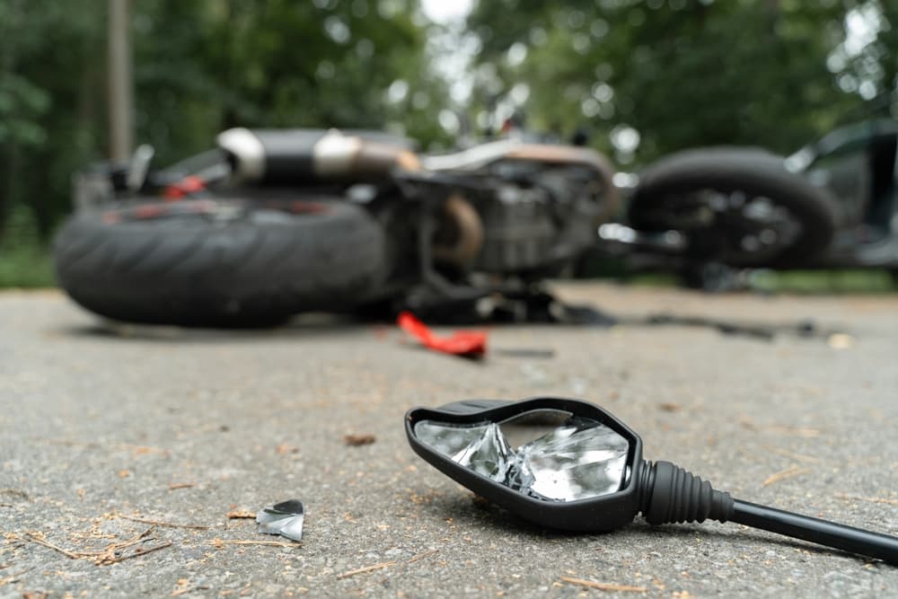 Broken motorcycle side mirror lying on the road with a damaged motorcycle in the background after an accident.
