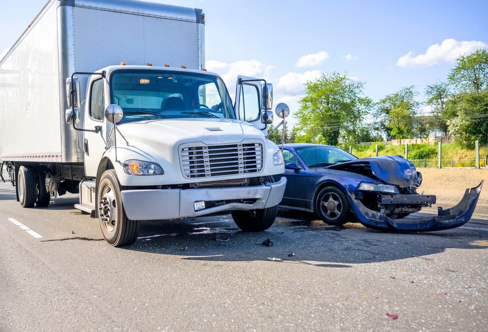 A damaged car and a large white truck after a collision on a sunny roadway, showing front-end impact and debris scattered on the road.