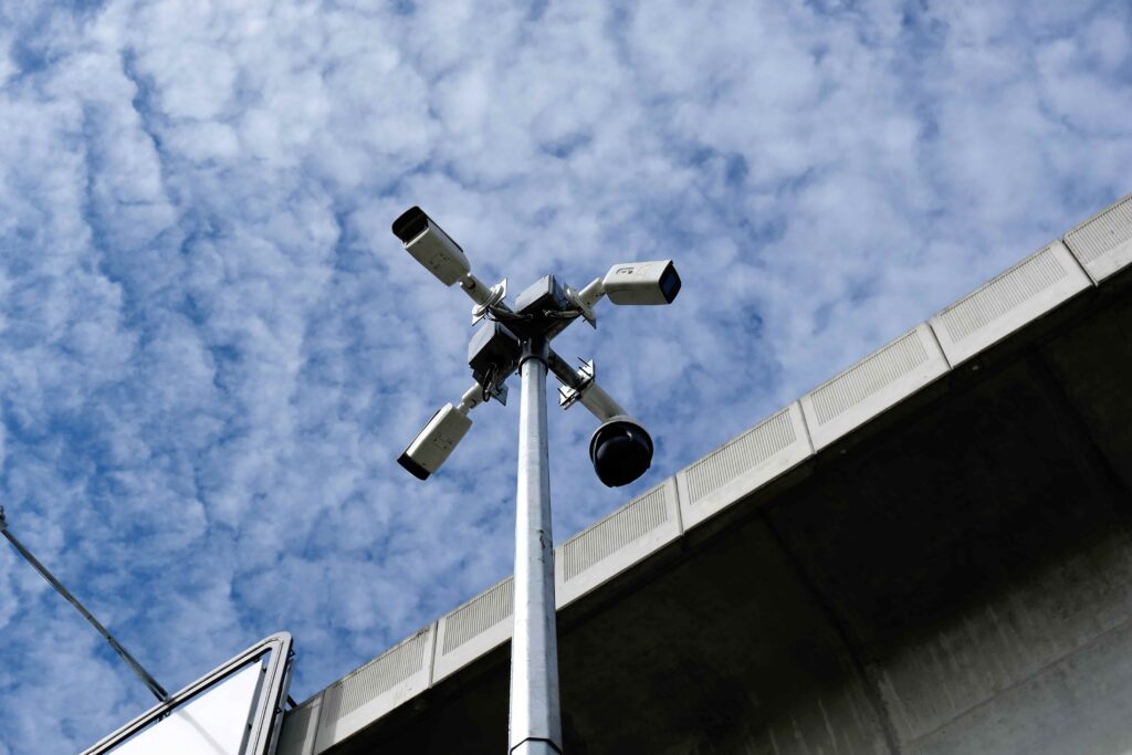Highway surveillance cameras mounted on a tall pole near an overpass, used for traffic monitoring and accident detection. Highway surveillance cameras mounted on a tall pole near an overpass, used for traffic monitoring and accident detection.