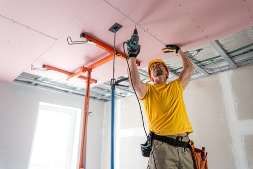 The worker screwing plasterboard to the ceiling. He is using special electric screwdriver and drywall lift. A red plasterboard improve the fire resistance of ceiling structures.