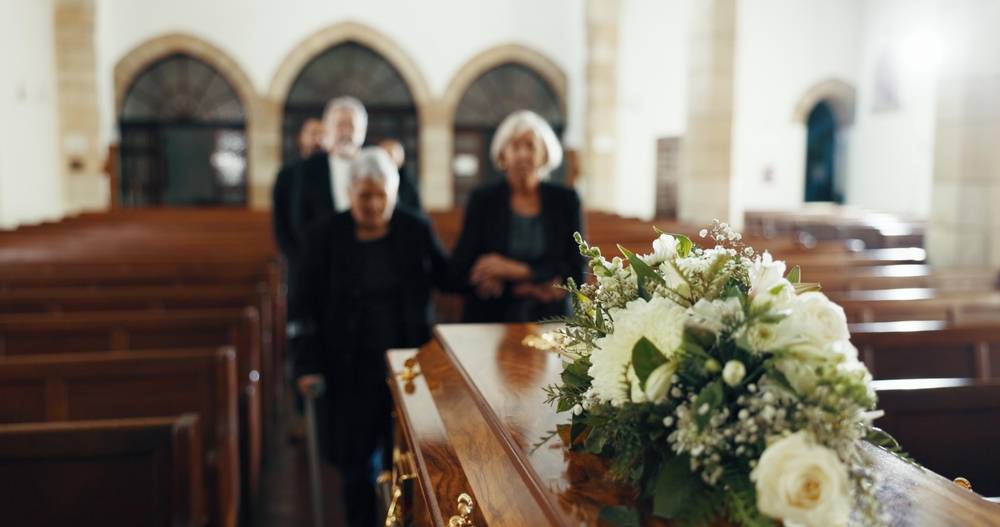 A casket decorated with white flowers in a church, with mourners in dark clothing walking towards it among wooden pews.