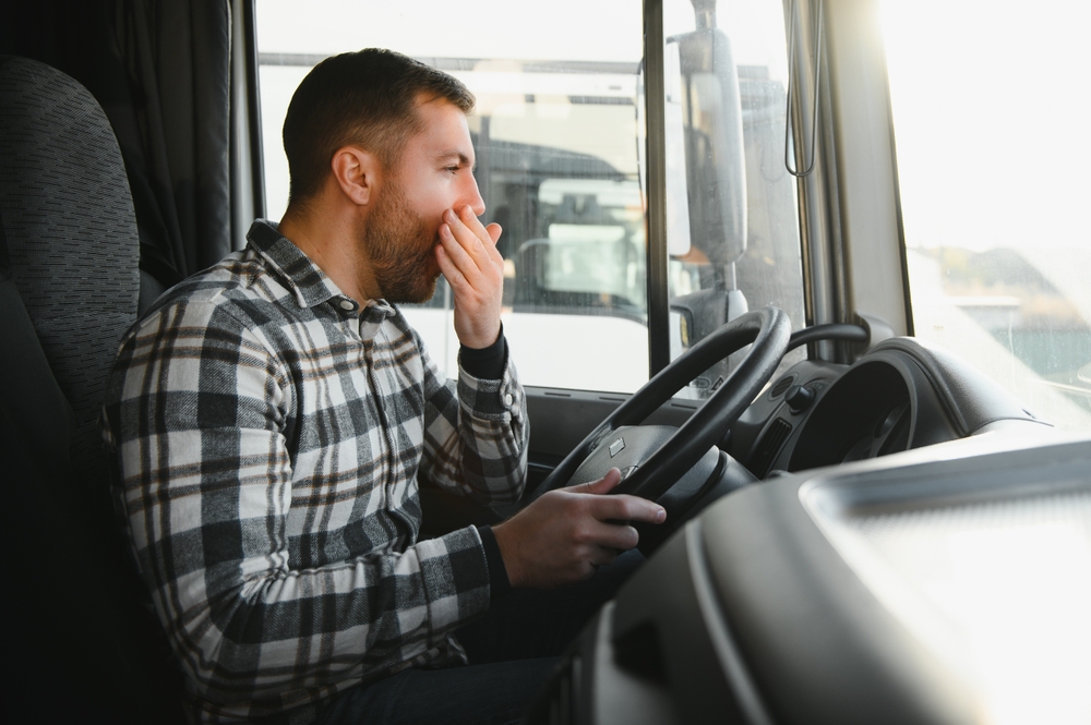 A man in a plaid shirt sitting in a truck, yawning while gripping the steering wheel.