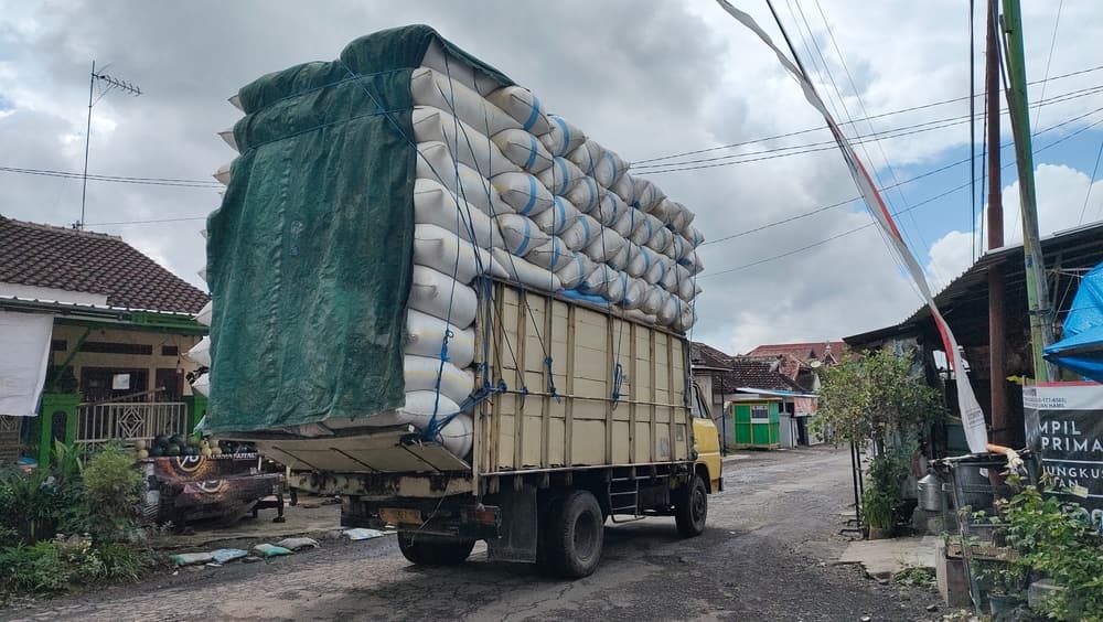A truck carrying a tall stack of bundled sacks traveling through a village street