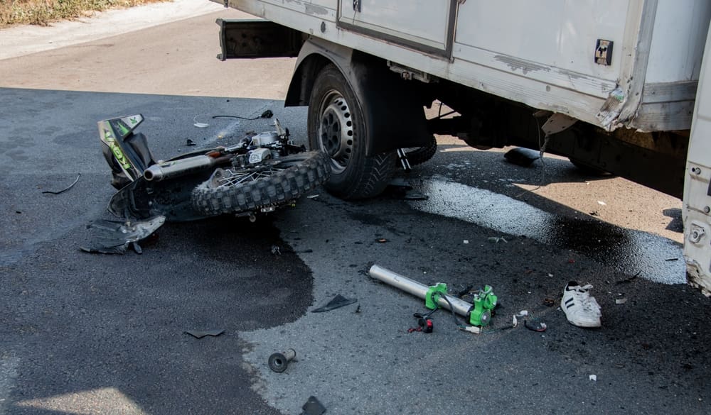 Damaged motorcycle trapped beneath a box truck with debris and spilled fluid scattered across the road